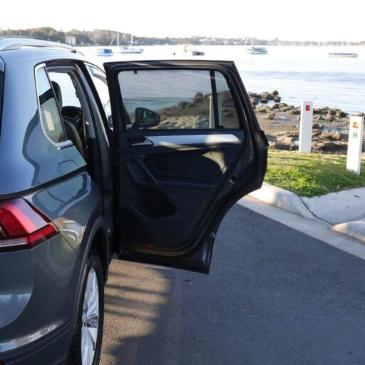 about-512x512 Open rear door of a gray SUV parked by a rocky waterfront with boats in the distance.